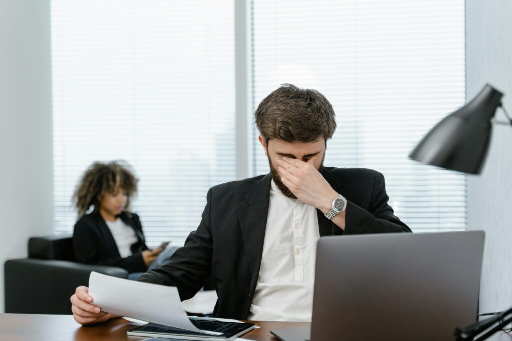 pexels-photo-7567197-7567197 Businessman in an office covering his eyes while working on a laptop, feeling stressed.